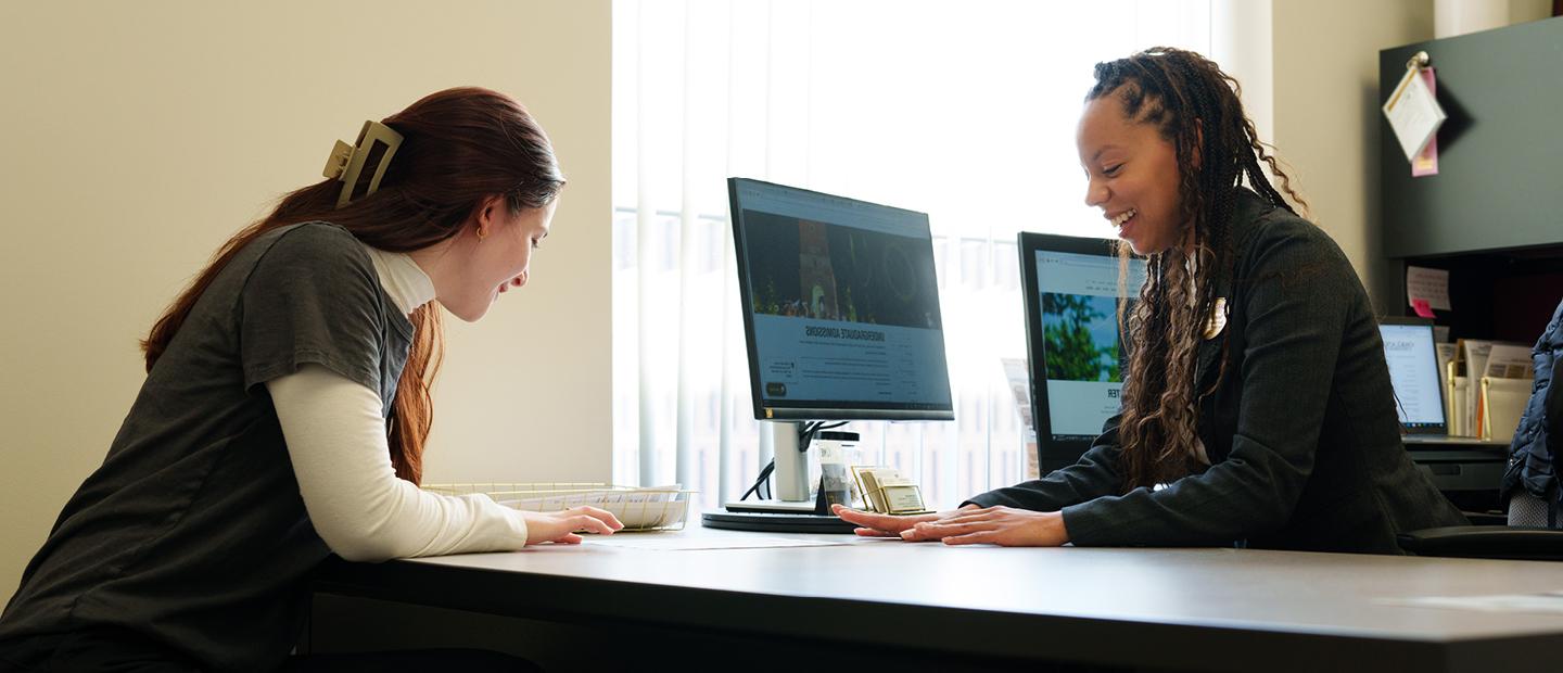 A woman looks at a document with another woman who sits across from her at a desk.